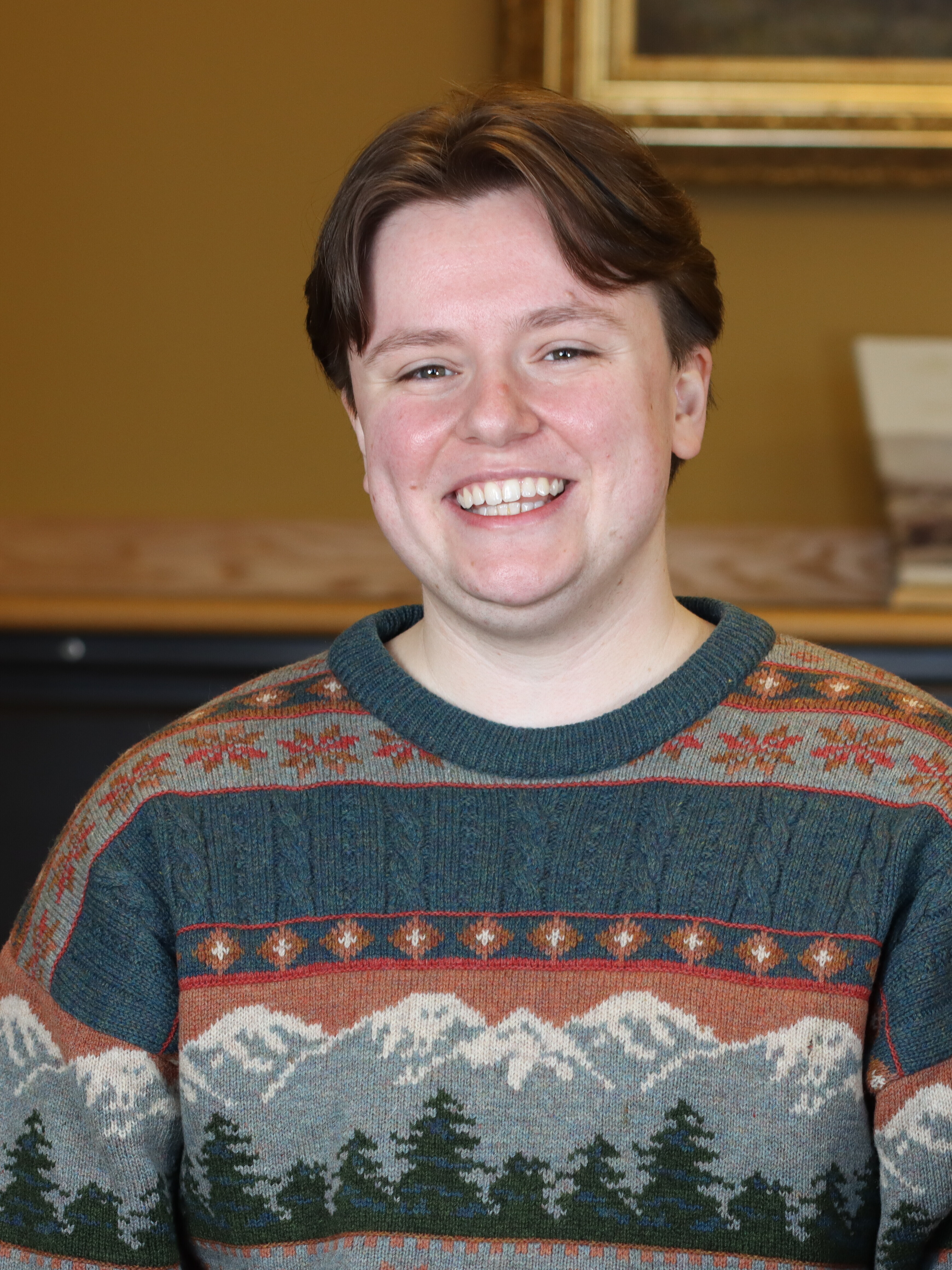 A person smiling at the camera. They're wearing a stripped sweater featuring mountains and trees.
