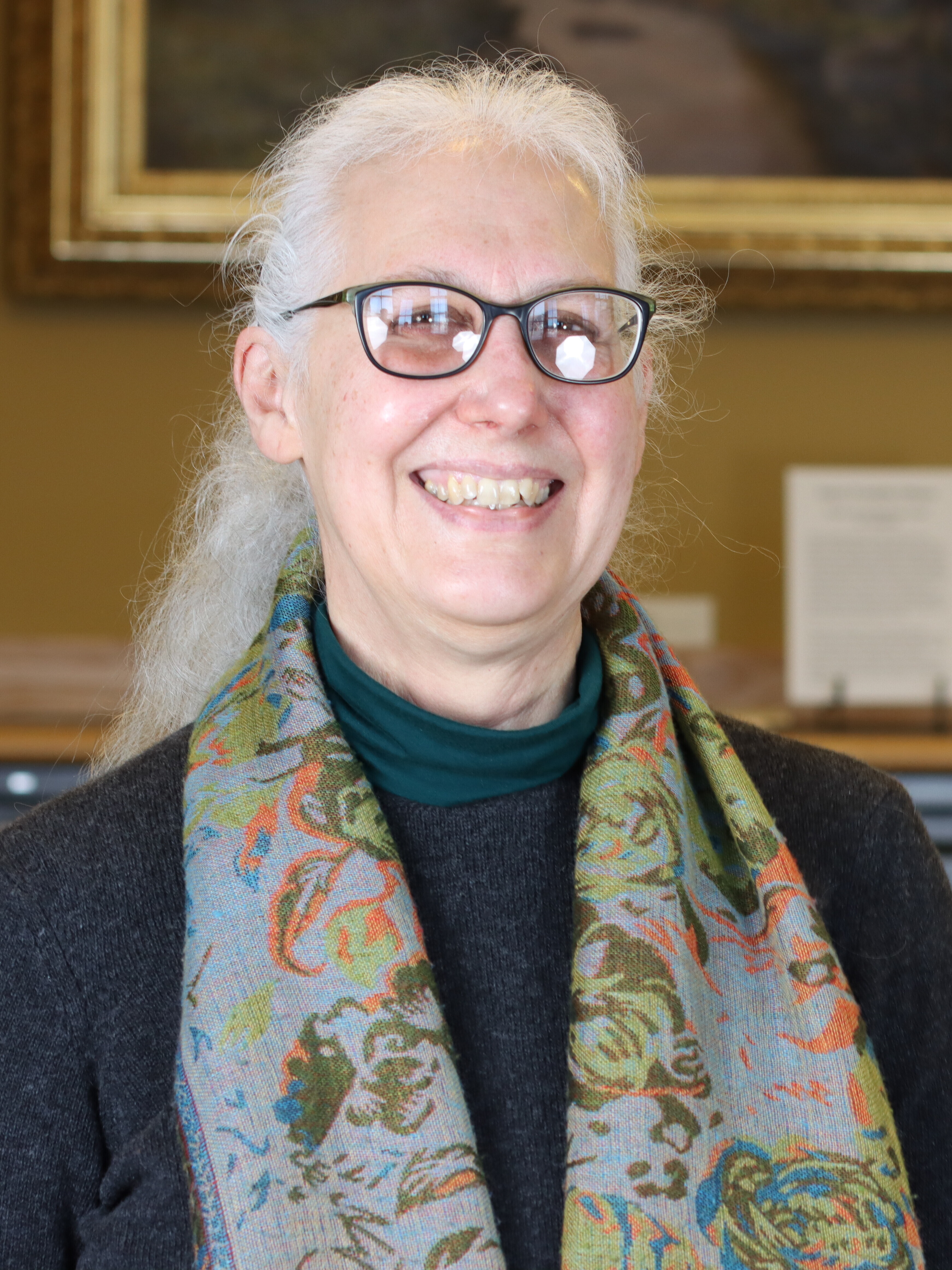 A white-haired woman with glasses smiles at the camera. She's wearing a black shirt with a red/orange/blue/green/gray scarf